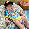 baby, child, outdoor, tie_dye, onesie, cap, hat, picnic, basket, cushion, striped, blanket, grass, smiling, happy, relaxed, infant, person, casual, colorful