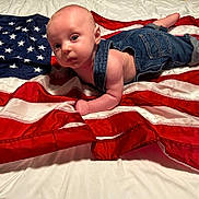Leo is registered to the contest to win money with this photo: baby, denim_overalls, american_flag, red, white, blue, infant, lying_down, curious, skin, fabric, patriotic, cute, clothing, portrait, child, indoors, soft_lighting, face, person