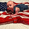 baby, denim_overalls, american_flag, red, white, blue, infant, lying_down, curious, skin, fabric, patriotic, cute, clothing, portrait, child, indoors, soft_lighting, face, person