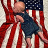 Leo is registered to the contest to win money with this photo: baby, denim_overalls, american_flag, infant, clothing, red_stripes, white_stripes, blue_field, stars, skin, barefoot, lying_down, portrait, child, cute, indoors, fabric, texture, soft_light, curious