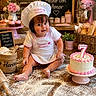child, toddler, baby, baker, chef_hat, apron, birthday_cake, number_seven, flour, baking, bakery, cupcakes, bread, pastries, kitchen, pink_mixer, roses, wooden_table, sitting, portrait