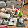 baby, smiling, toy, play_mat, cushion, couch, blanket, floor, child, indoor, happy, person, plush_toy, head, footwear, clothing, face, home, tiled_floor, seated