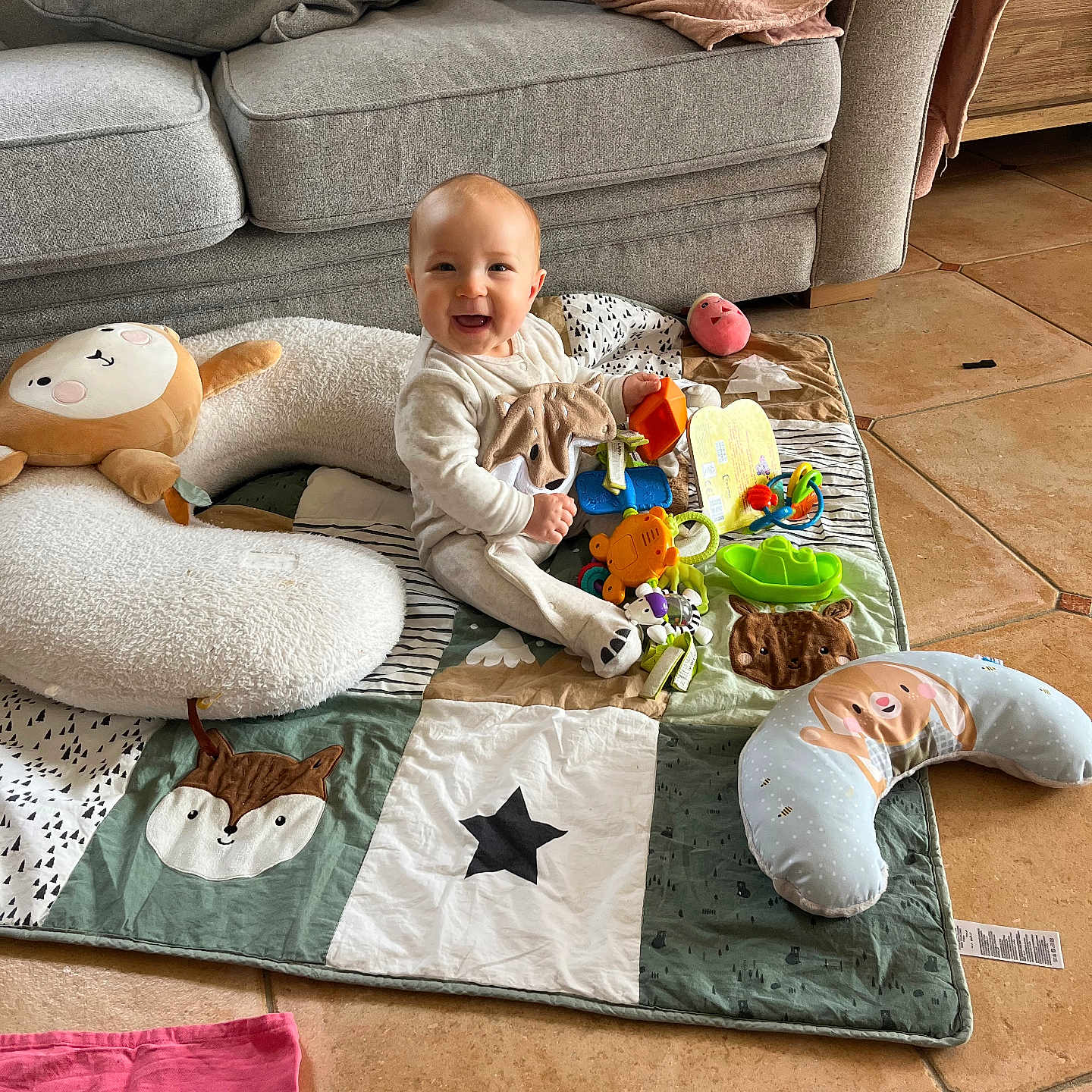 Lyam a rejoint le concours — aidez-le/la à gagner de superbes lots ! baby, smiling, toy, play_mat, cushion, couch, blanket, floor, child, indoor, happy, person, plush_toy, head, footwear, clothing, face, home, tiled_floor, seated