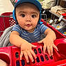 baby, child, shopping_cart, blue_cap, striped_shirt, store_aisle, textiles, shelves, curious, hands, indoor, retail, shopping, infant, person, cute, wide_eyes, red, clothing, baby_in_cart