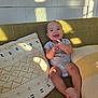 baby, smile, couch, pillow, indoor, child, happy, sunlight, patterned_pillow, sitting, infant, clothing, barefoot, face, cute, relaxing, home, cozy, young_child, joyful