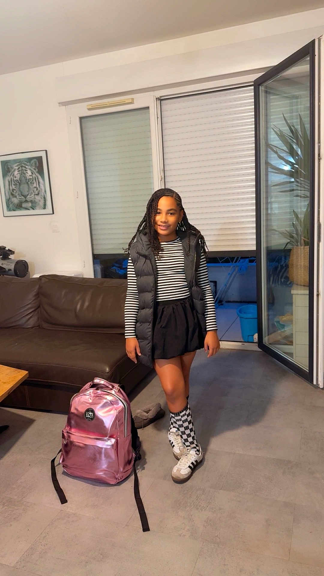 Ivy participe au concours pour gagner de l'argent avec cette photo : child, girl, backpack, living_room, couch, window, open_door, plant, sneakers, checkered_socks, striped_shirt, puffer_vest, skirt, braids, portrait, smile, floor, painting, indoor, home