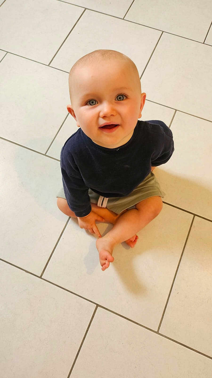 Jules participe au concours pour gagner de l'argent avec cette photo : toddler, child, baby, floor, tile, sitting, cross_legged, smile, blue_eyes, short_hair, casual_clothing, long_sleeve, shorts, indoor, cute, looking_up, barefoot, young_child, curious, person