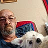 bald, beard, casual_clothing, companion, couch, dog, framed_picture, furry_pet, glasses, home, indoor, man, pet, pillow, portrait, relaxing, senior, snuggling, wall_art, white_dog