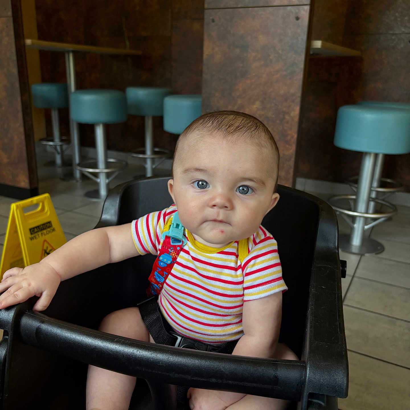 Daylan is registered to the contest to win money with this photo: baby, child, high_chair, striped_shirt, blue_eyes, indoor, restaurant, bar_stools, yellow_sign, wet_floor_sign, curious, seated, black_chair, floor_tiles, pacifier_clip, colorful_clothing, infant, person, cute, casual