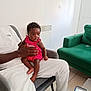 baby, child, adult, indoor, living_room, chair, couch, phone, screen, clothing, sitting, person, hand, footwear, tile_floor, curly_hair, pink_outfit, white_pants, green_sofa, table