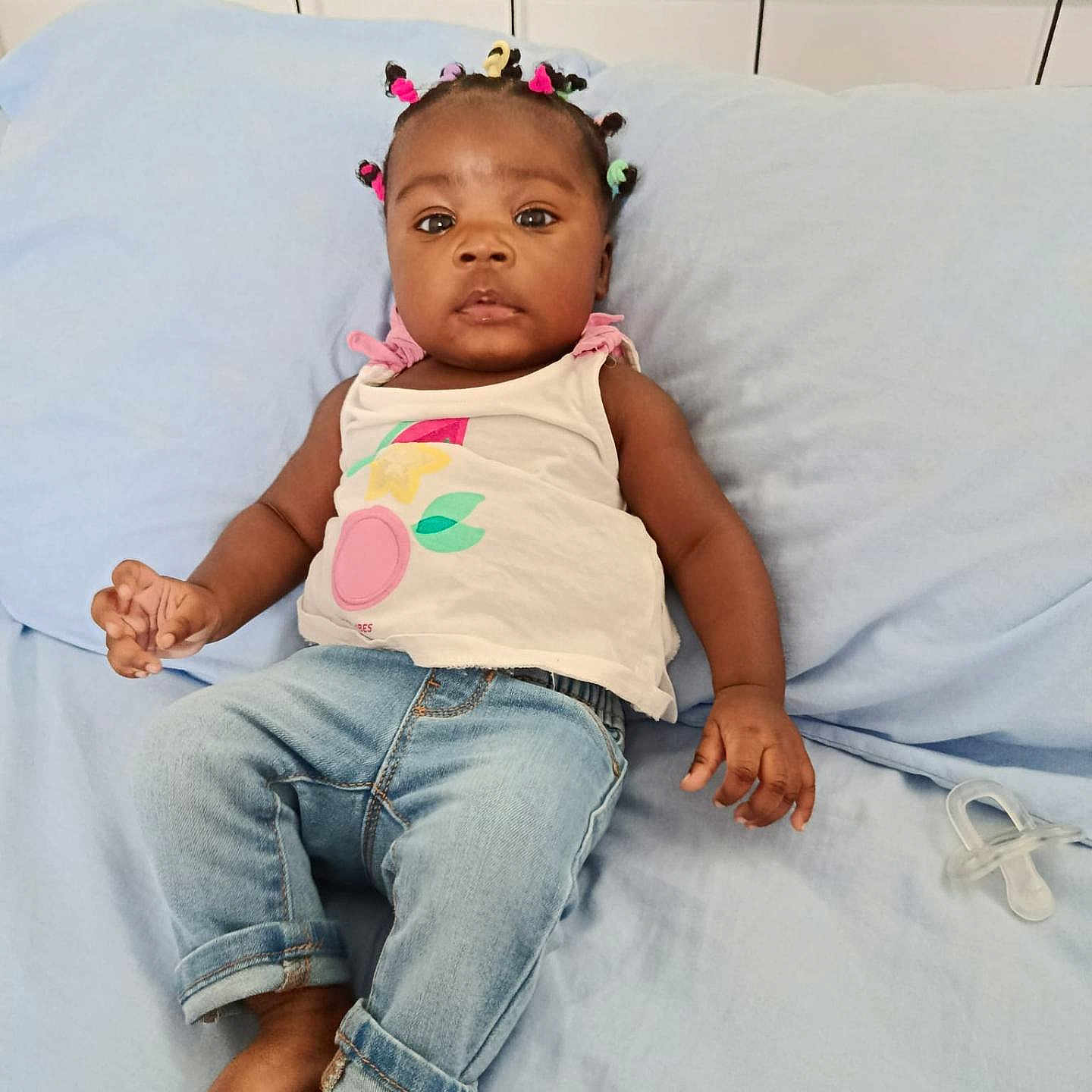 Maelys Kelssie participe au concours pour gagner de l'argent avec cette photo : baby, bare_feet, bed, blue_bedspread, casual_clothing, child, colorful_hair_ties, cute, face, hand, indoors, infant, jeans, pacifier, person, portrait, relaxed, skin, white_top, young_child