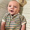 baby, infant, child, blue_eyes, striped_shirt, colorful_clothing, plaid_blanket, wooden_sign, milestone, portrait, smiling, happy, cute, lying_down, close_up, face, skin, short_hair, indoors, young_child