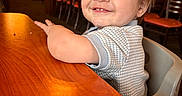 Sebastian joined the competition — help win amazing prizes! toddler, child, high_chair, smiling, table, wood, restaurant, indoor, happy, person, face, clothing, hair, seat, light, background_blur, expression, cute, portrait, furniture