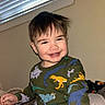 toddler, child, smiling, indoor, dinosaur_shirt, clothing, face, hair, window_blind, light, expression, person, young_child, cute, happy, casual, portrait, sitting, playful, room