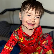 Sebastian is registered to the contest to win money with this photo: baby, bed, bright_colors, child, clothing_pattern, cute, face, hair, happy, head, home, indoor, pajamas, person, portrait, red_clothing, sitting, smiling, toddler, young_child