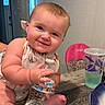 Avery is registered to the contest to win money with this photo: baby, child, smiling, water_bottle, table, high_chair, indoor, face, person, cute, happy, short_hair, apparel, beads, drinks, plastic_cup, food_container, foot, hand, expression