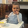 bowl, chair, child, clothing, denim_overalls, eating, face, floor, food, happy, high_chair, indoor, long_sleeve_shirt, person, plant, restaurant, smiling, table, toddler, wall
