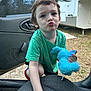 child, toddler, green_shirt, stuffed_toy, blue_toy, pacifier, car_door, car_seat, outdoor, grass, camper, playful, pout, young_child, person, short_hair, daylight, casual, cute, toy
