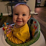 Paislee is registered to the contest to win money with this photo: baby, child, infant, smiling, happy, portrait, indoor, living_room, activity_seat, high_chair, yellow_clothing, toy, playtime, television, wood_floor, home, sitting, face, hands, colorful