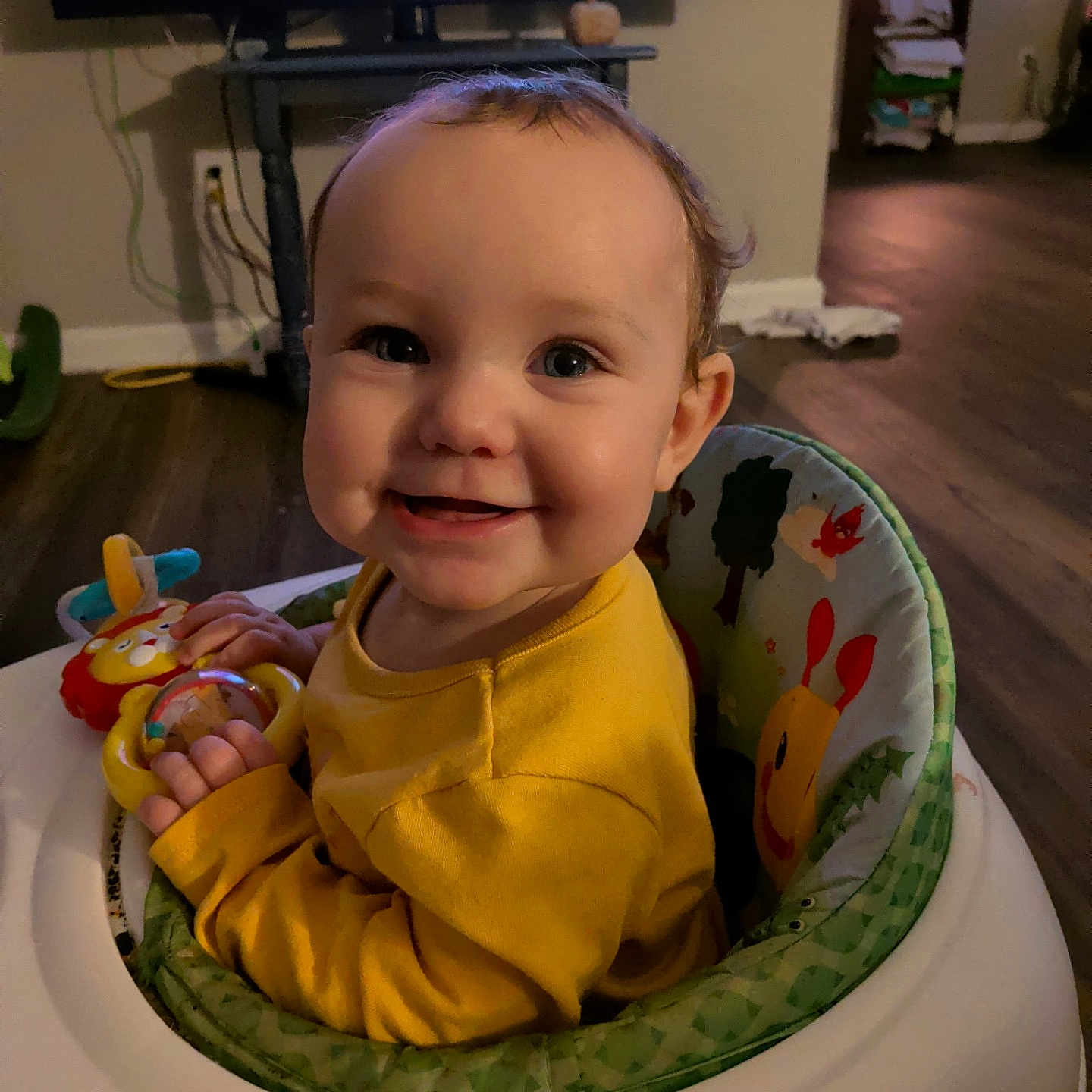 Paislee is registered to the contest to win money with this photo: baby, child, infant, smiling, happy, portrait, indoor, living_room, activity_seat, high_chair, yellow_clothing, toy, playtime, television, wood_floor, home, sitting, face, hands, colorful