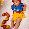 apples, baby, blanket, blue_top, bokeh, child, colorful, cute, fluffy, fruit, indoors, infant, lights, lying_down, portrait, princess_outfit, red_headband, smiling, soft_texture, yellow_bottom