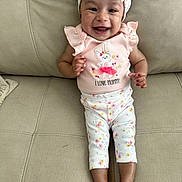 Eliana is registered to the contest to win money with this photo: baby, infant, smiling, headband, flower_pattern, pink_clothing, bunny_design, text_on_clothing, sitting, couch, beige_furniture, happy, cute, child, person, indoors, legs, barefoot, portrait, young