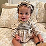 baby, child, smile, curly_hair, headband, dress, patterned_clothing, sitting, couch, pillows, indoors, footwear, cute, happy, portrait, infant, person, home, soft_lighting, fashion
