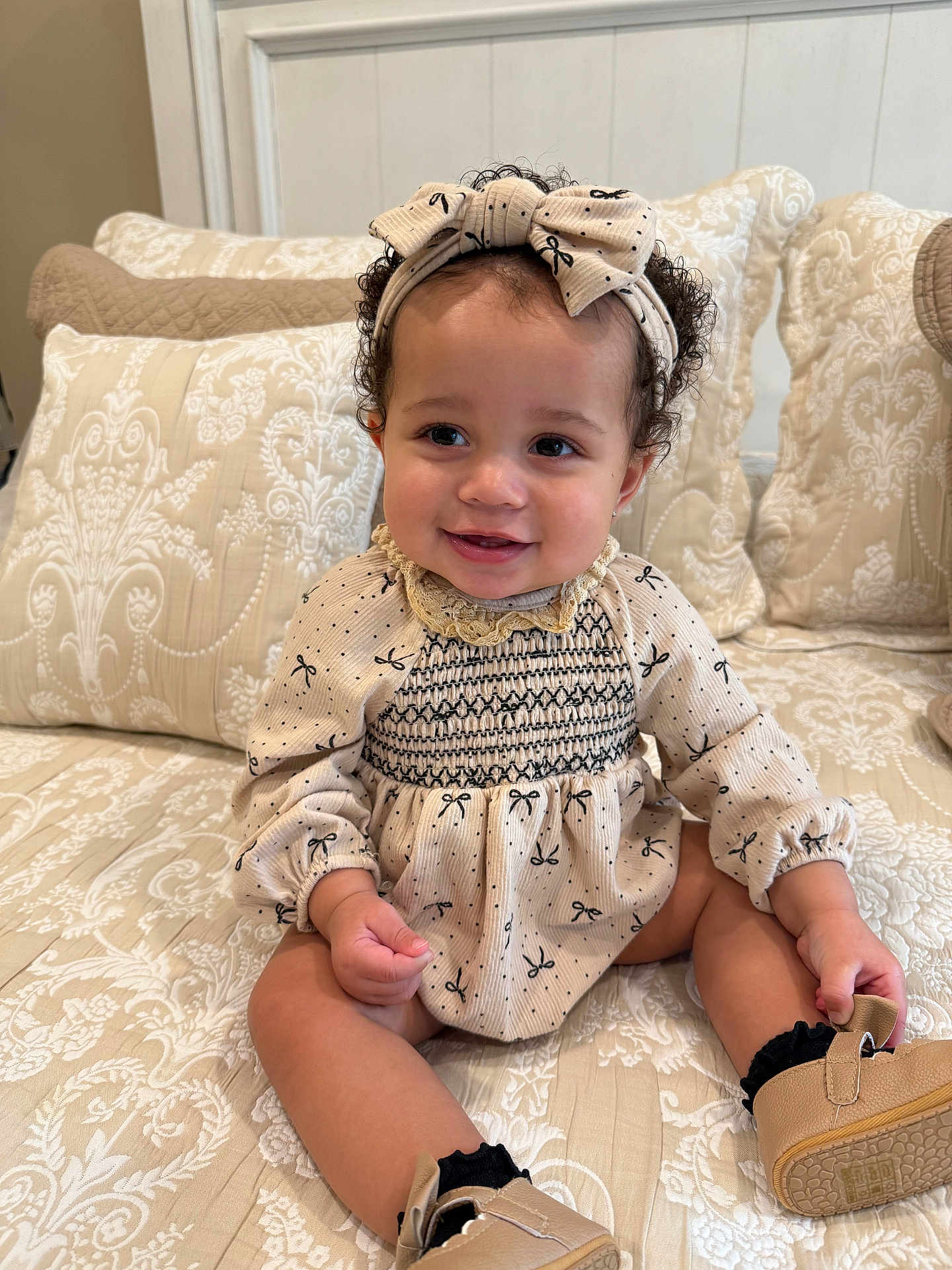 Brielle joined the competition — help win amazing prizes! baby, child, smile, curly_hair, headband, dress, patterned_clothing, sitting, couch, pillows, indoors, footwear, cute, happy, portrait, infant, person, home, soft_lighting, fashion