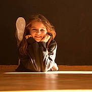 Éléna participe au concours pour gagner de l'argent avec cette photo : child, smiling, floor, wooden_floor, long_hair, casual_clothing, indoors, warm_light, happy, person, face, sneakers, black_sweater, resting, relaxed, portrait, young, natural_light, cozy, casual