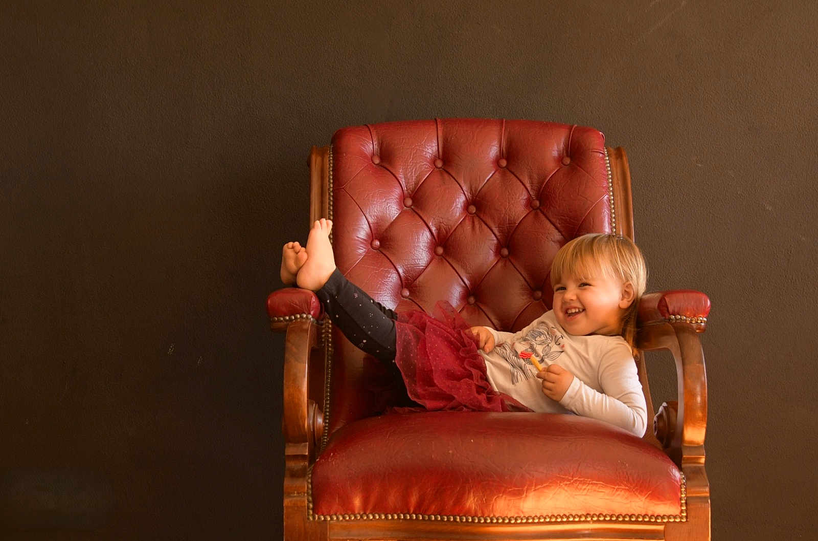 Ayana participe au concours pour gagner de l'argent avec cette photo : child, toddler, smiling, chair, red_leather, armchair, legs_up, long_sleeve_shirt, skirt, indoor, wooden_armrests, happy, blond_hair, casual_clothing, relaxed, furniture, portrait, cute, person, home