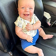 William is registered to the contest to win money with this photo: baby, child, smiling, sitting, car_seat, black_leather, clothing, shorts, shirt, mickey_mouse, happy, cute, infant, skin, face, hand, foot, seat, interior, vehicle