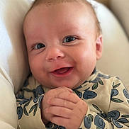 Wilfred is registered to the contest to win money with this photo: baby, smiling, hands, blueberry_pattern, clothing, face, infant, cute, happy, skin, eyes, mouth, person, portrait, closeup, sitting, indoors, cushion, comfort, young_child