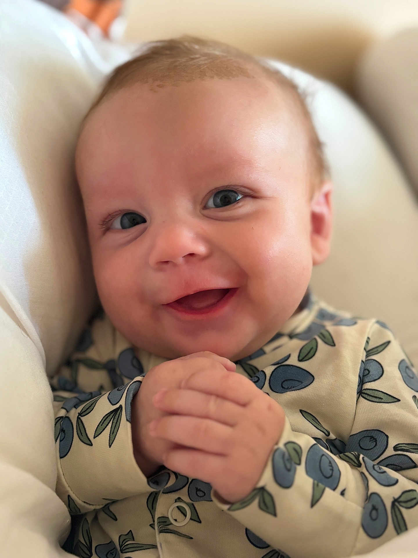 Wilfred is registered to the contest to win money with this photo: baby, smiling, hands, blueberry_pattern, clothing, face, infant, cute, happy, skin, eyes, mouth, person, portrait, closeup, sitting, indoors, cushion, comfort, young_child