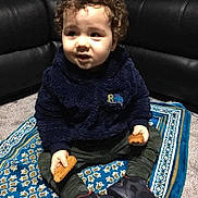 Hunter joined the competition — help win amazing prizes! child, toddler, curly_hair, cookie, rug, blue_rug, fleece_jacket, navy_fleece, pants, sneakers, black_couch, indoor, carpet, sitting, holding_food, curious_expression, person, young_child, casual_clothing, home