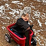 toddler, child, wagon, radio_flyer, hat, knitted_hat, bow, outdoor, ground, dirt, white_debris, red, black_jacket, toy, wheels, handle, sitting, cold_weather, expression, person