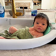 Liam is registered to the contest to win money with this photo: baby, bath, bathtub, bubbles, towel, water, sink, kitchen, countertop, faucet, cleaning, child, infant, wet_hair, household, soap, blue_bottle, pink_cap, relaxing, curious