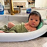 baby, bath, bathtub, bubbles, towel, water, sink, kitchen, countertop, faucet, cleaning, child, infant, wet_hair, household, soap, blue_bottle, pink_cap, relaxing, curious