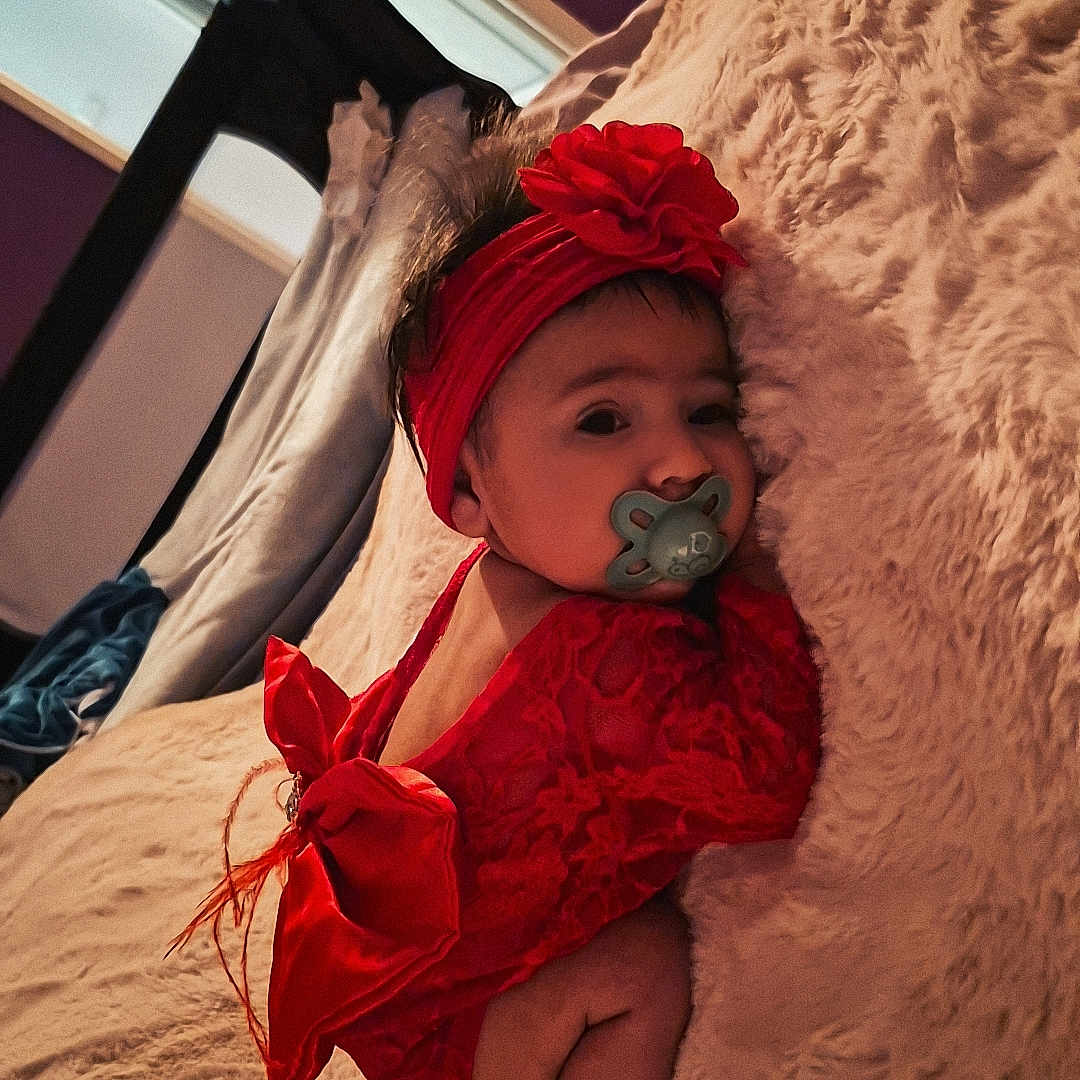 Zénaya participe au concours pour gagner de l'argent avec cette photo : baby, bed, blanket, child, cozy, curious, cute, flower, headband, indoor, infant, lying_down, pacifier, person, portrait, purple_wall, red_clothing, small_feet, soft_texture, window