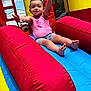 toddler, child, smiling, pink_tank_top, shorts, inflatable_slide, red, yellow, blue, outdoor, sunny, fun, play, happy, arms_up, barefoot, summer, daytime, excited, recreation