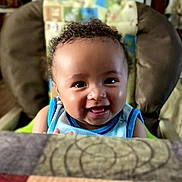 Riley is registered to the contest to win money with this photo: baby, child, smiling, curly_hair, high_chair, indoor, portrait, happy, cute, infant, person, face, clothing, blue, seat, expression, young, cheerful, skin, head