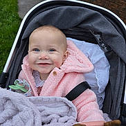 Luna is registered to the contest to win money with this photo: baby, stroller, pink_coat, blanket, toy, smile, blue_eyes, outdoor, grass, sidewalk, happy, child, face, clothing, person, portrait, infant, warm_clothing, cute, cozy