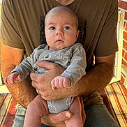 Kyrin is registered to the contest to win money with this photo: baby, infant, person, adult, hand, clothing, onesie, elephant_print, blue_eyes, face, legs, arms, shirt, jeans, chair, cushion, brick_wall, outdoor, seated, holding