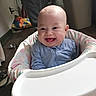 baby, high_chair, smiling, happy, tongue_out, blue_clothing, indoor, person, floor, furniture, home, child, infant, seat, playful, cute, expression, human, babysuit, joy