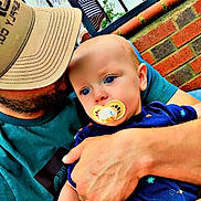 Walt joined the competition — help win amazing prizes! baby, pacifier, man, cap, arm, holding, blue_eyes, brick_wall, fence, outdoor, people, clothing, hand, closeup, child, person, casual, daylight, portrait, tender_moment