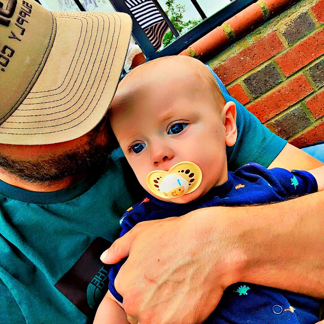 Walt joined the competition — help win amazing prizes! arm, baby, blue_eyes, brick_wall, cap, casual, child, closeup, clothing, daylight, fence, hand, holding, man, outdoor, pacifier, people, person, portrait, tender_moment