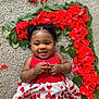 baby, smiling, red_rose_petals, heart_shape, carpet, red_top, white_skirt, red_hearts, curly_hair, cute, infant, happy, flower_arrangement, green_leaves, lying_down, portrait, child, adorable, indoor, closeup