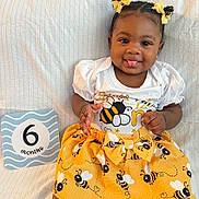 Layla is registered to the contest to win money with this photo: baby, infant, child, smiling, tongue_out, bee_pattern, yellow_skirt, white_shirt, hair_bows, cute, playful, sitting, blanket, sign, 6_months, portrait, closeup, happy, indoors, person
