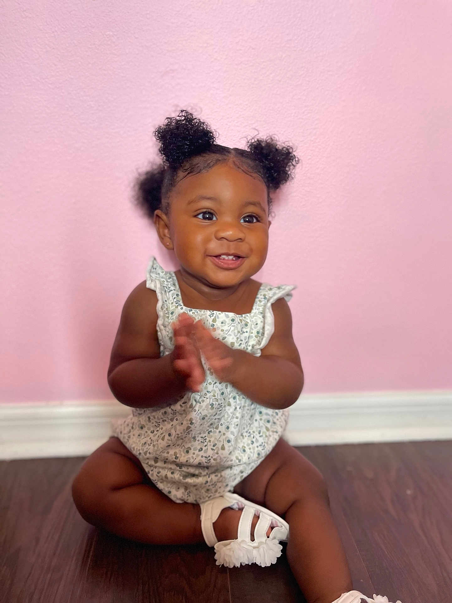 Layla is registered to the contest to win money with this photo: baby, child, smiling, clapping, curly_hair, pigtails, sitting, floor, pink_wall, white_dress, white_sandals, indoors, cute, happy, portrait, person, infant, skin, footwear, wooden_floor