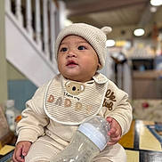 Aiden joined the competition — help win amazing prizes! baby, infant, bottle, milk, hat, knit_hat, bib, clothing, sitting, indoor, table, face, cute, child, person, milk_bottle, warm_lighting, cozy, soft_focus, curious