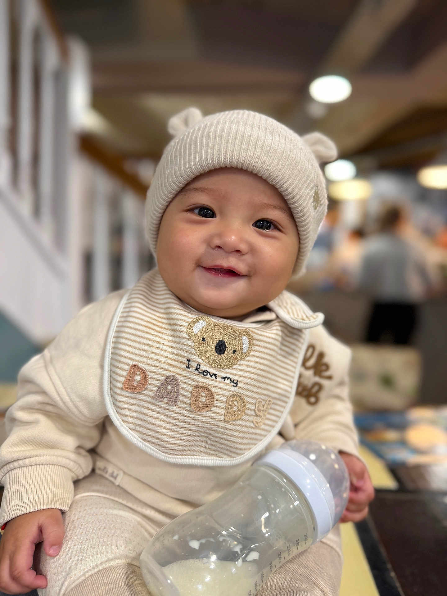 Aiden joined the competition — help win amazing prizes! baby, infant, smiling, bottle, hat, beige_clothing, bib, indoor, cute, child, portrait, happy, milk, feeding, warm_clothing, face, person, cozy, seated, closeup